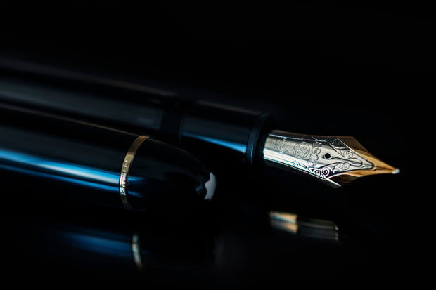 Ein Montblanc Meisterstück-Füllfederhalter mit goldener 14-Karat-Feder liegt geöffnet auf schwarzem Untergrund. Die glänzende Kappe mit goldener Zierleiste und dem weißen Montblanc-Emblem ist sichtbar im Vordergrund.